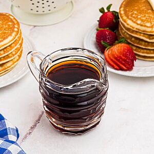 Homemade syrup in a glass pitcher in front of stacks of pancakes with strawberries.
