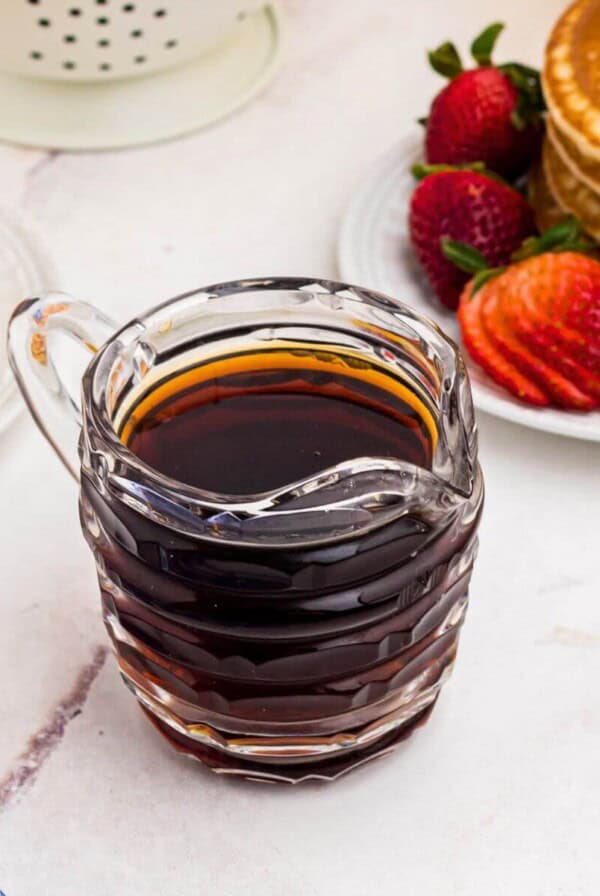 Homemade syrup in a glass pitcher in front of stacks of pancakes with strawberries.