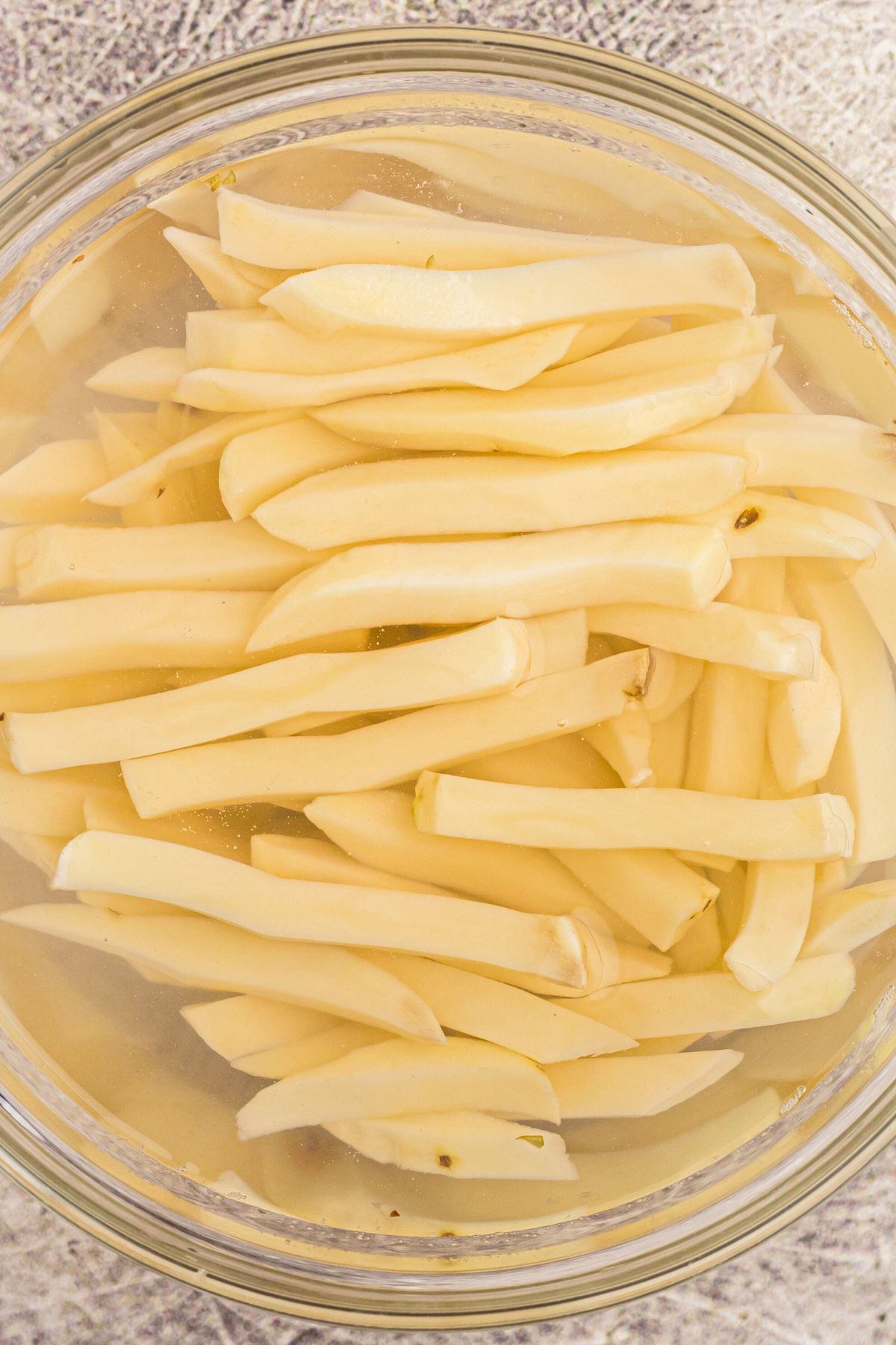 Potato slices cut into fries, being soaked in a clear glass bowl filled with cold water. 