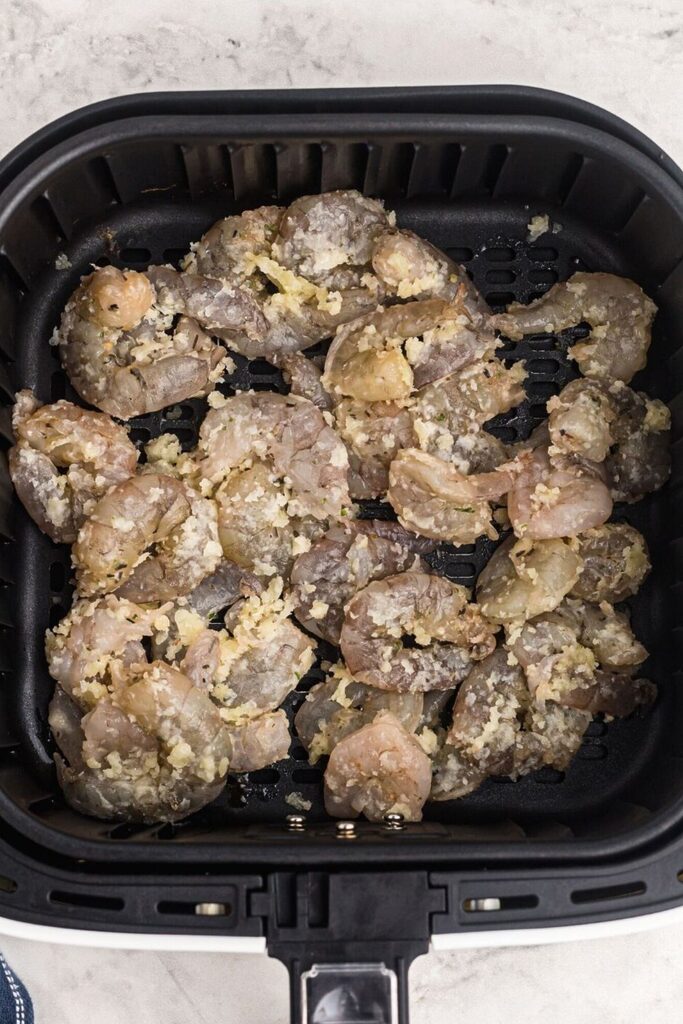 Raw and uncooked shrimp in the air fryer basket, coated with butter and garlic. 