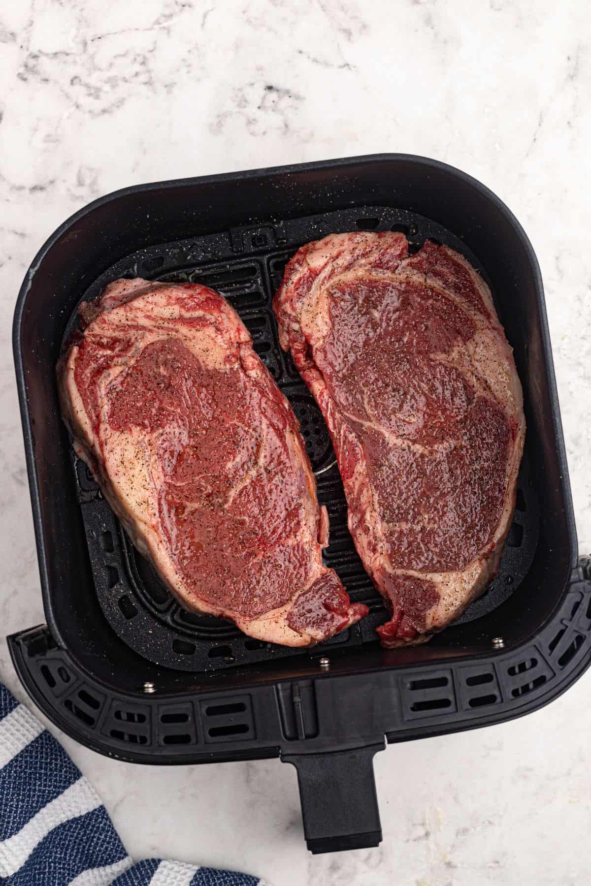 Uncooked seasoned steaks in the air fryer basket before being cooked on a marble table. 