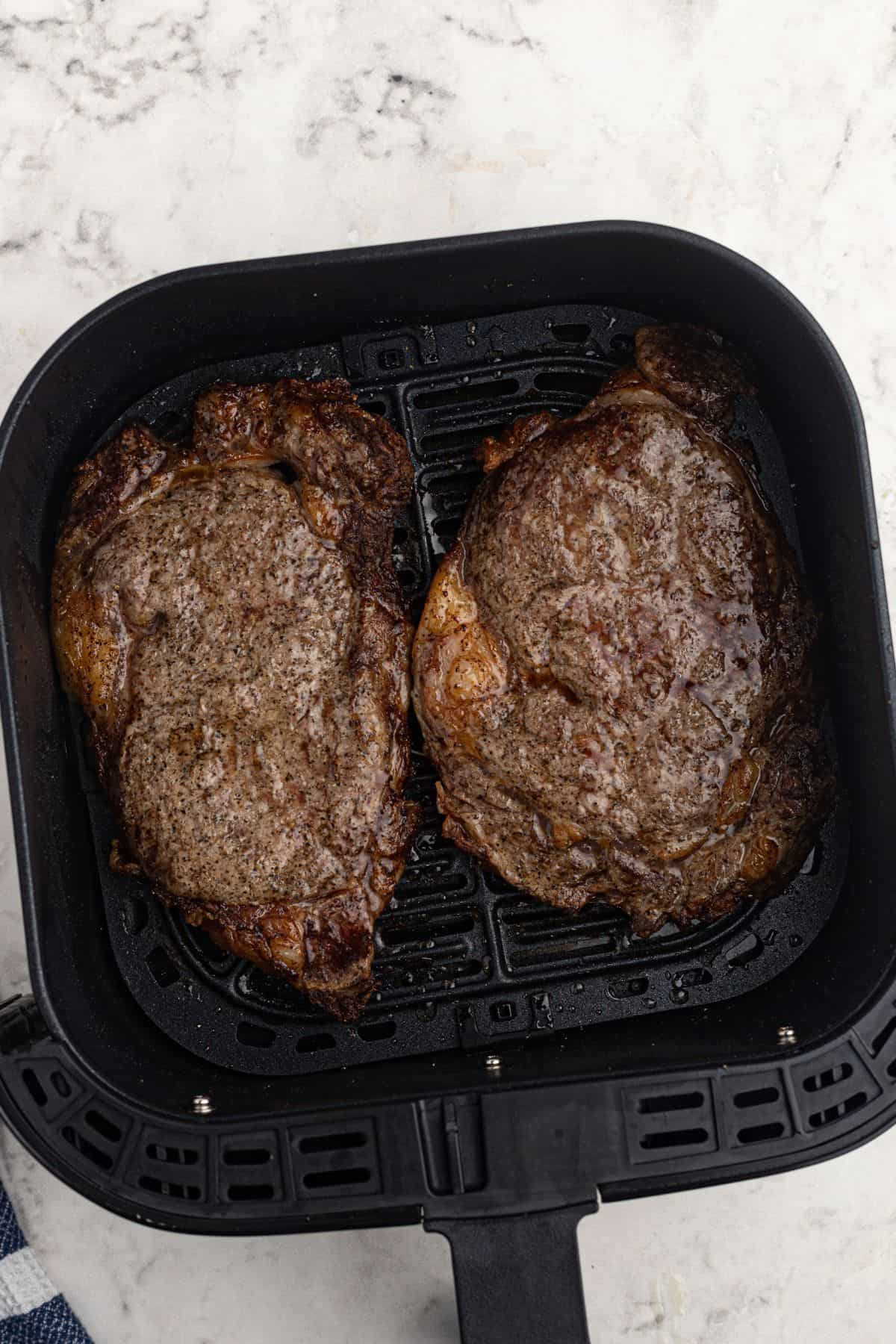 Two juicy cooked ribeye steaks in the air fryer basket after being seasoned and cooked. 