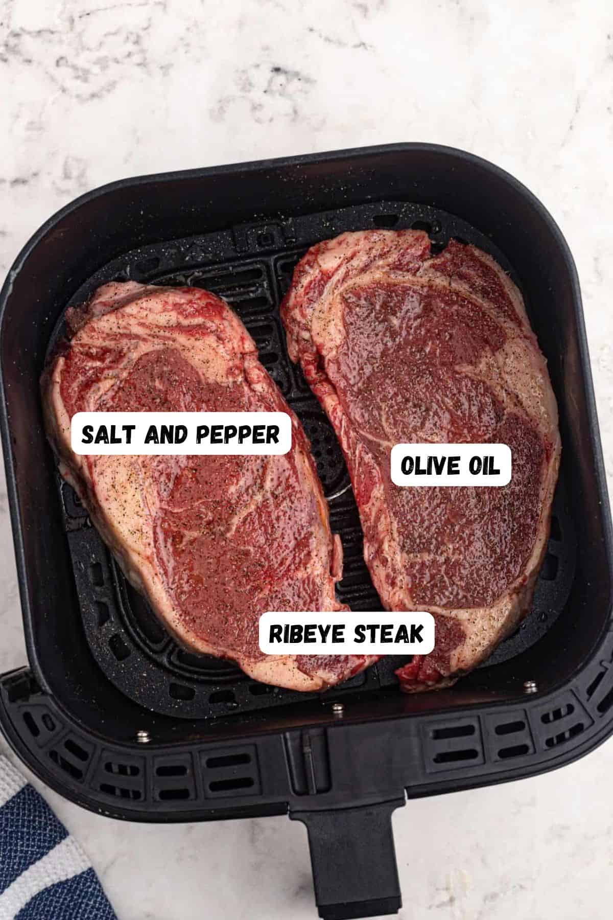 Uncooked ribeye steaks in an air fryer basket with olive oil, salt, and pepper. 