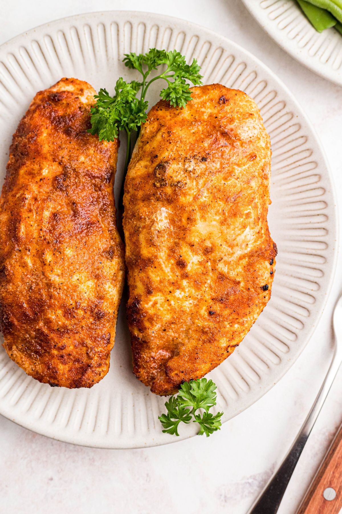 Two seasoned golden chicken breasts on a white plate with parsley garnish. 