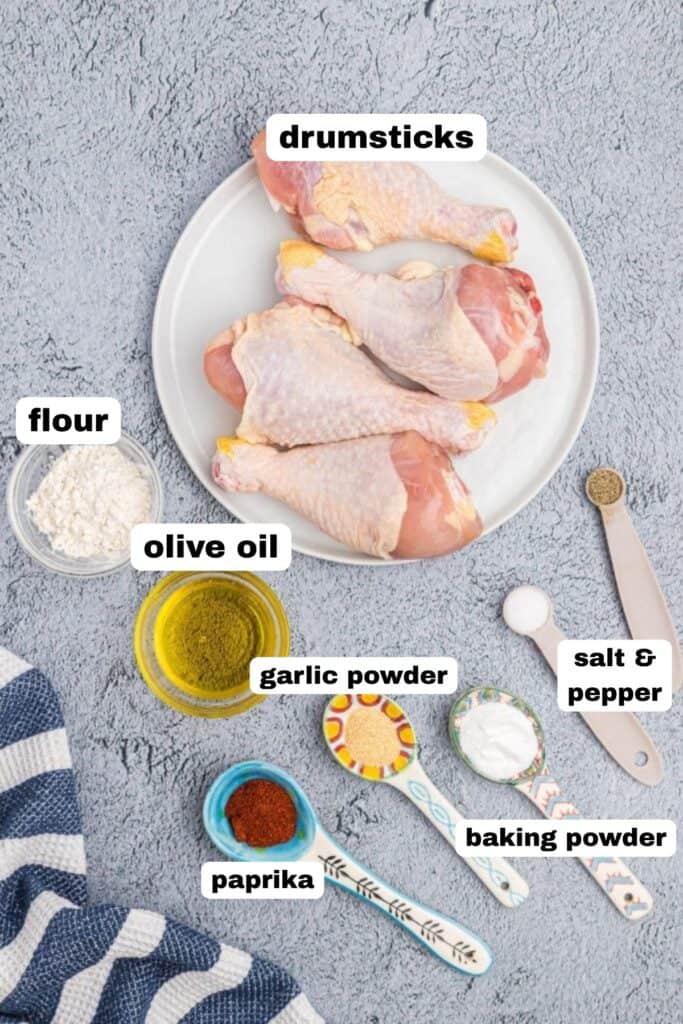 Ingredients needed to make drumsticks, seasonings, olive oil, and uncooked chicken, in dishes and measuring spoons, on a light gray table. 