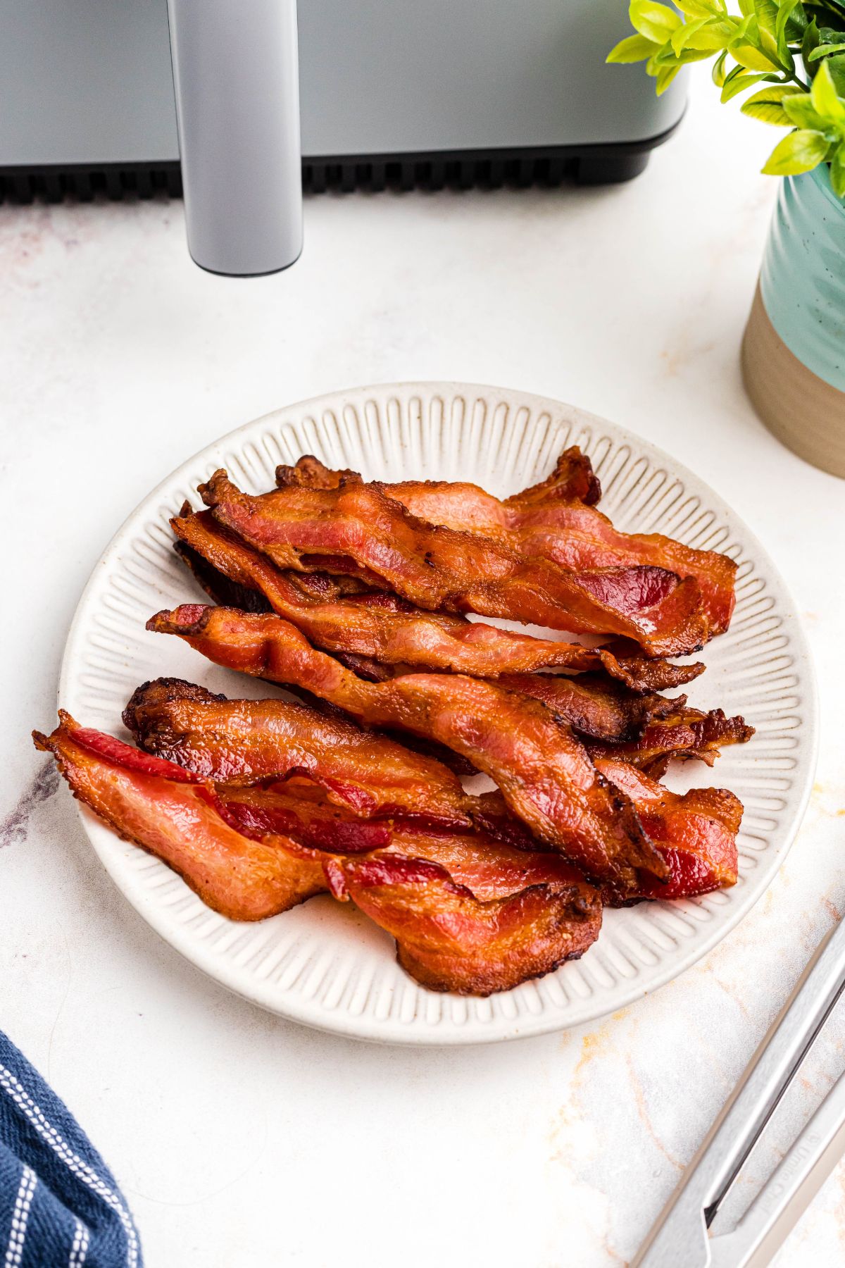 Golden crispy bacon stacked on a cream colored plate in front of a gray air fryer. 