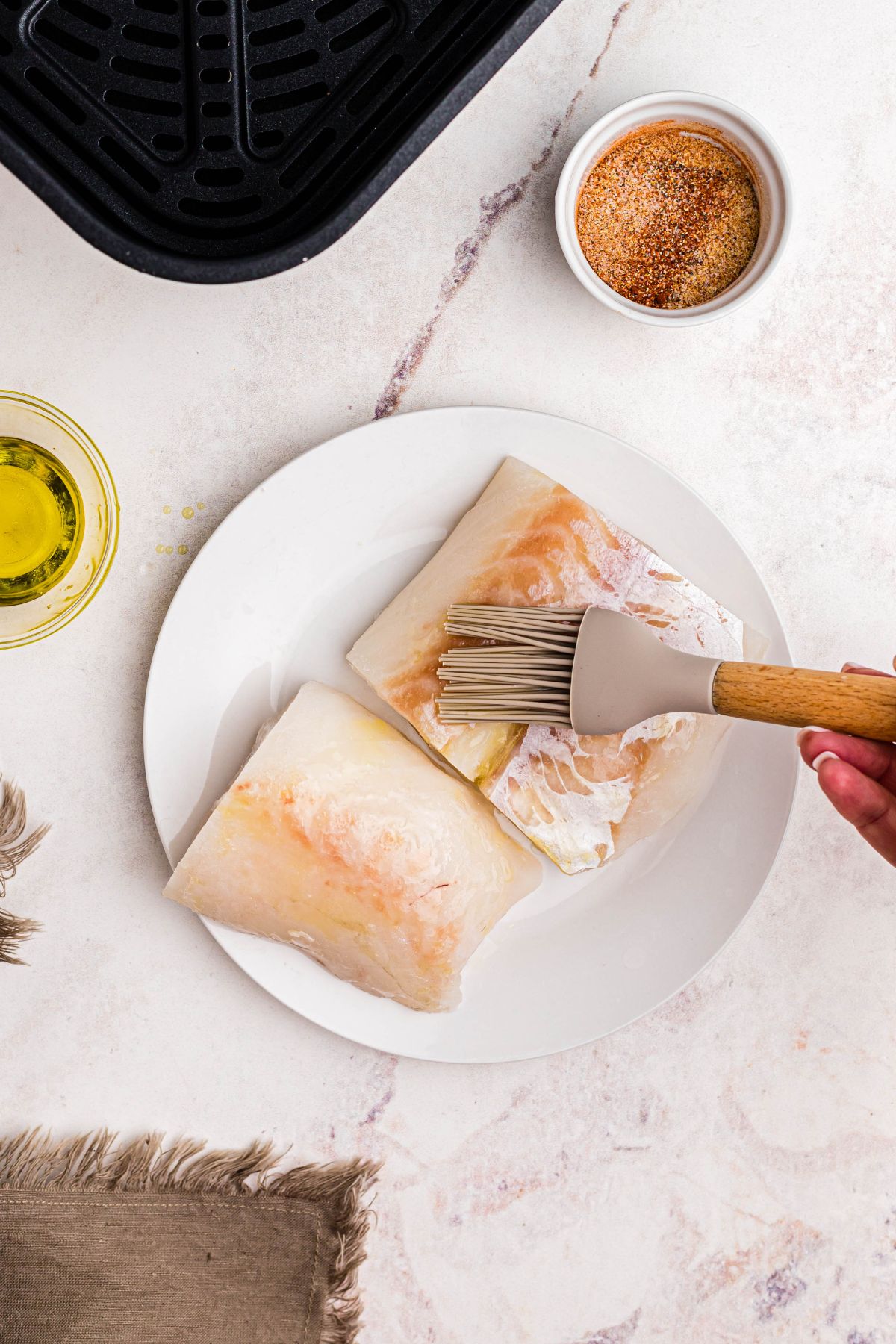 Brushing olive oil on frozen cod filets on a white plate before air frying. 