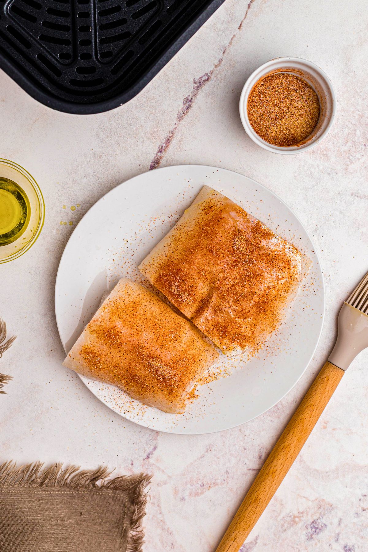 Seasoned frozen cod on a white plate in front of the air fryer basket. 