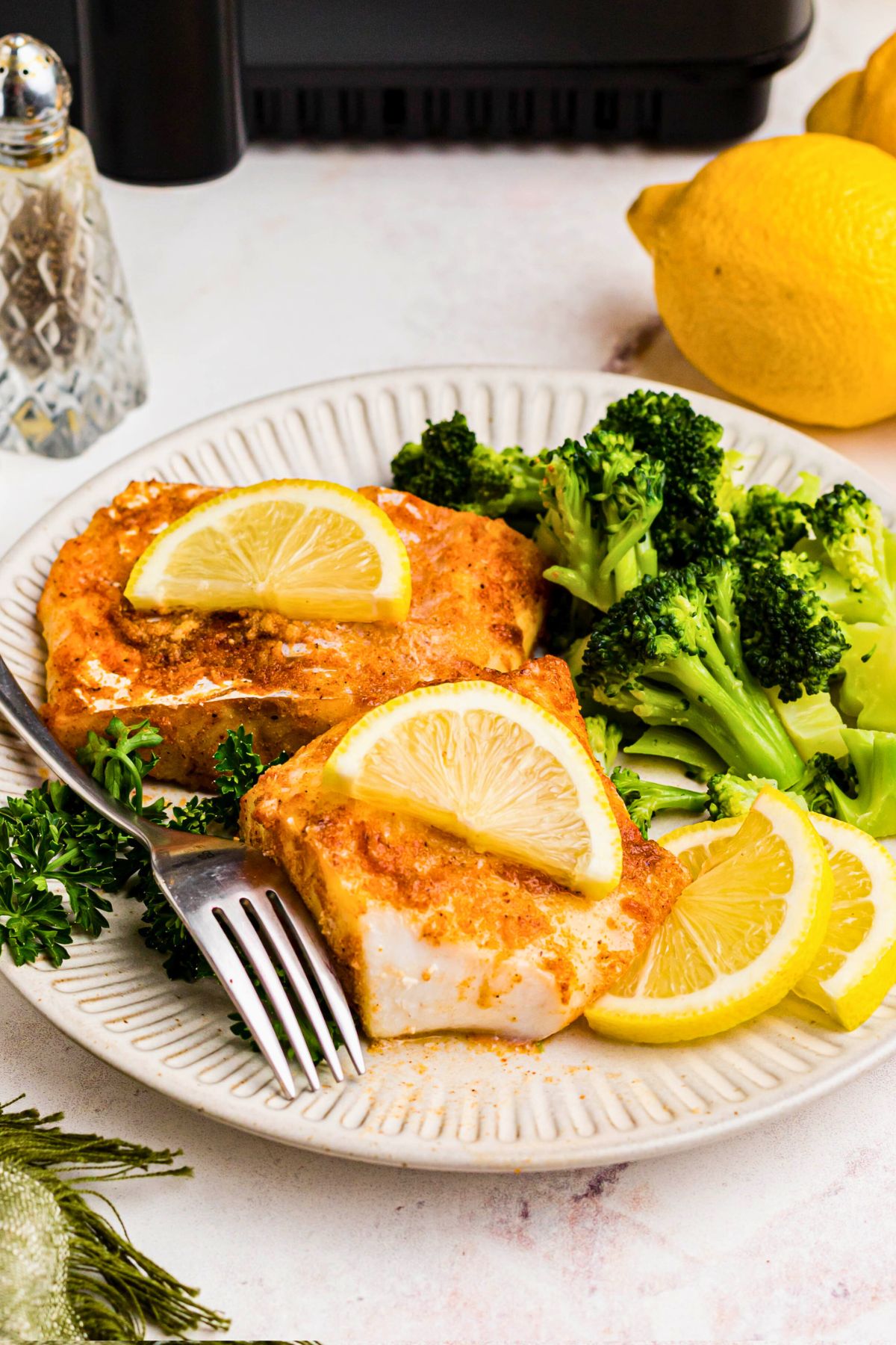 Cooked and seasoned cod on a white plate with broccoli and lemon slices, with a fork laying across the plate. 