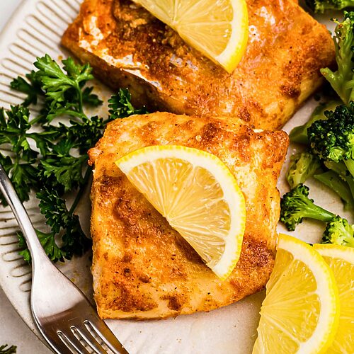 Juicy seasoned cod on a white plate, served with broccoli and lemon slices.