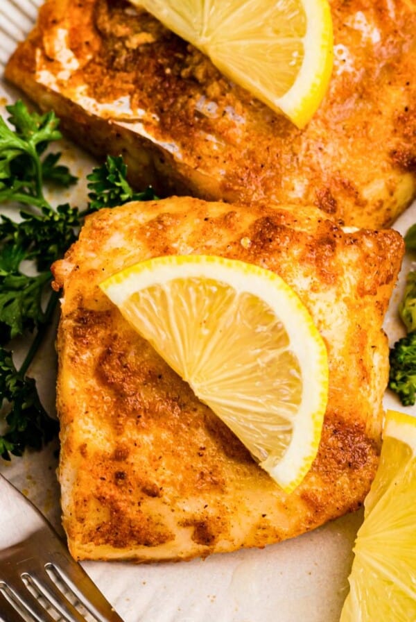 Juicy seasoned cod on a white plate, served with broccoli and lemon slices.