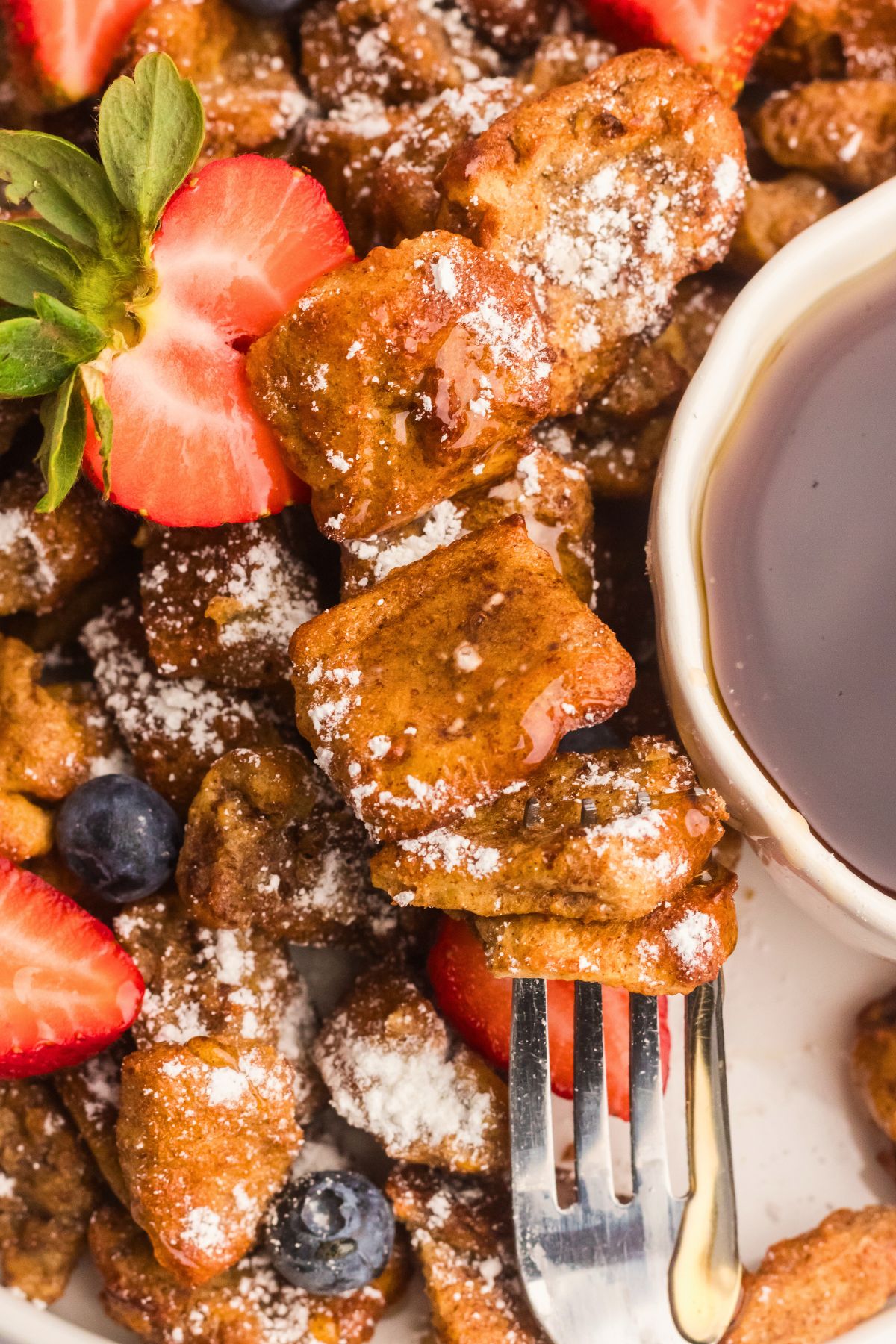 Golden French toast bites on a plate with a few bites on a fork with syrup and strawberries. 