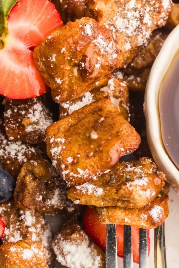 Golden French toast bites on a white plate with berries and syrup.