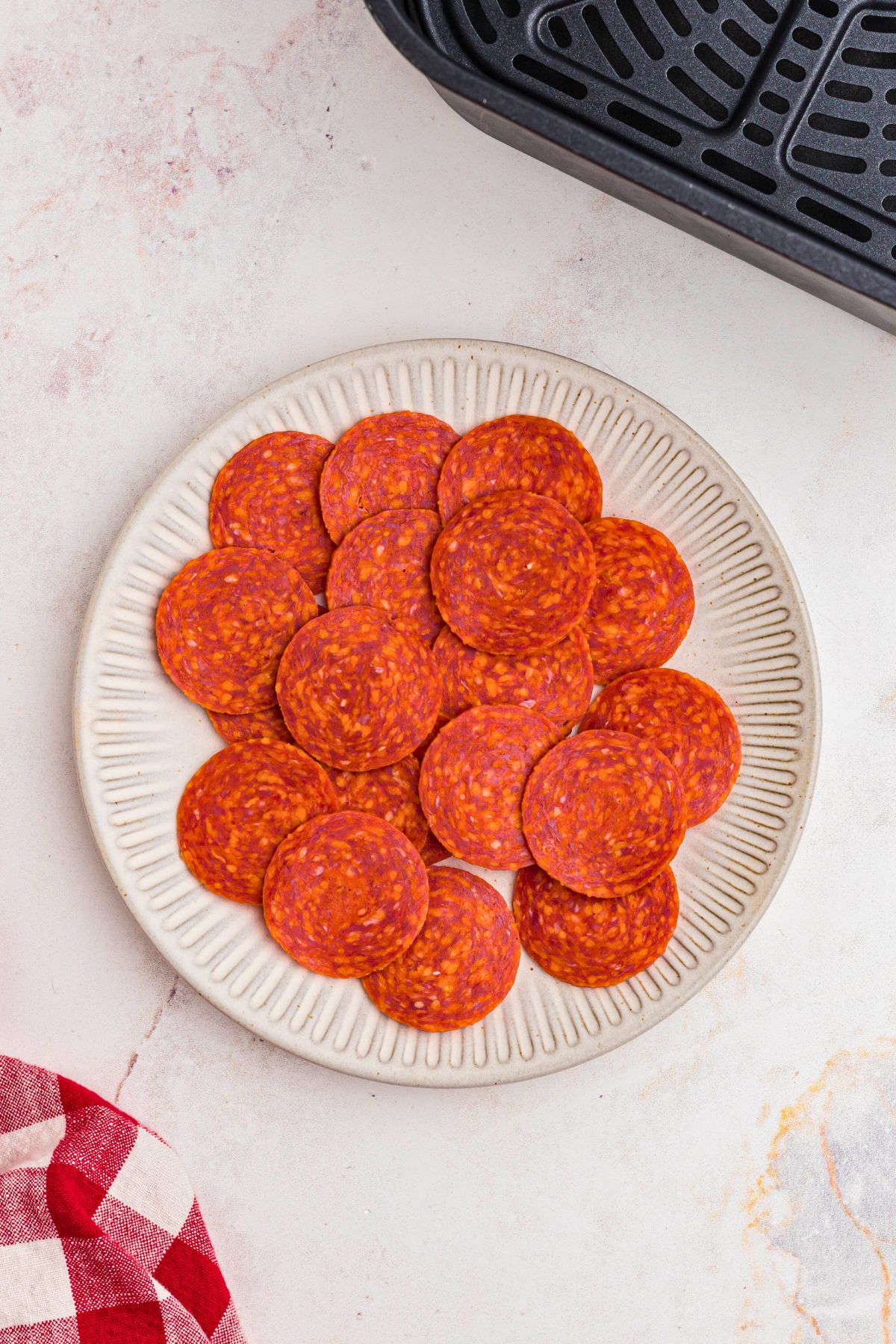 Uncooked slices of pepperoni on a round white plate in front of an air fryer. 