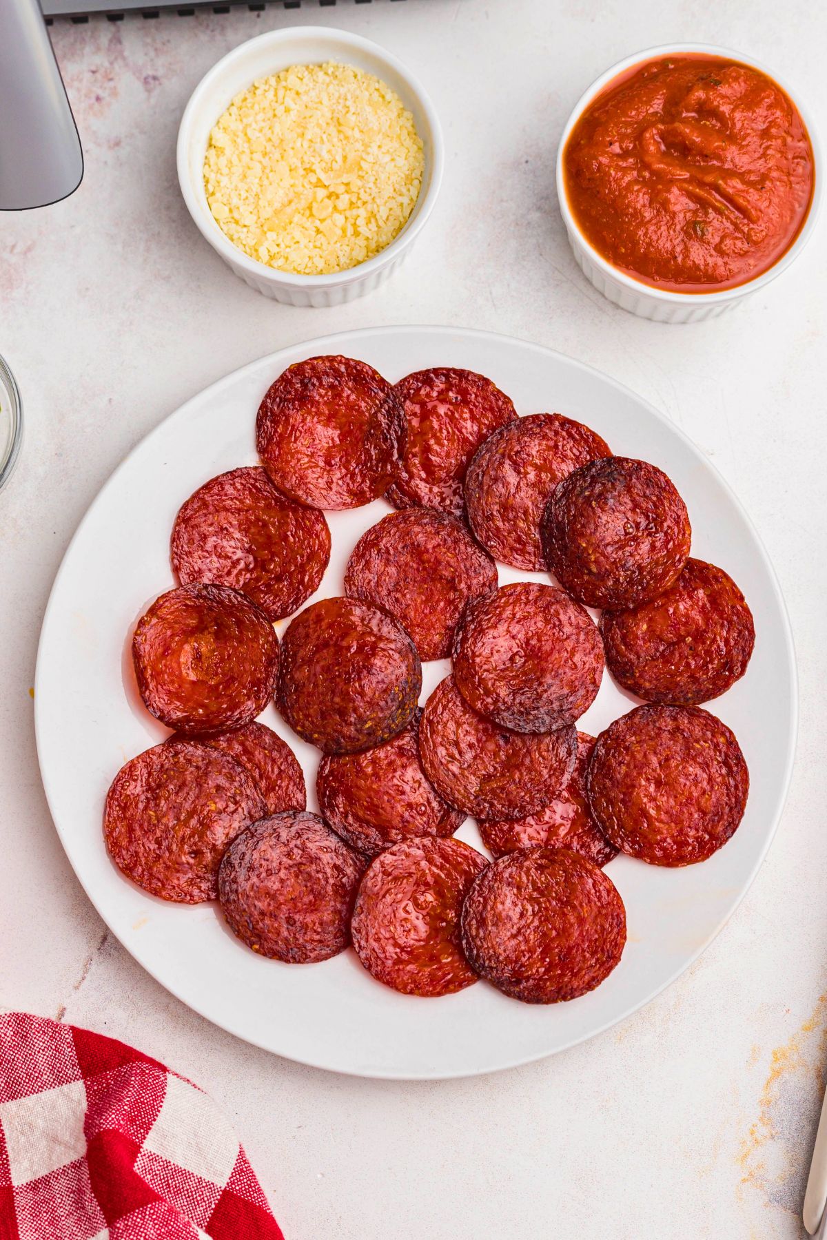 Red pepperoni chips on a white plate with pizza sauce and parmesan cheese in small dishes. 