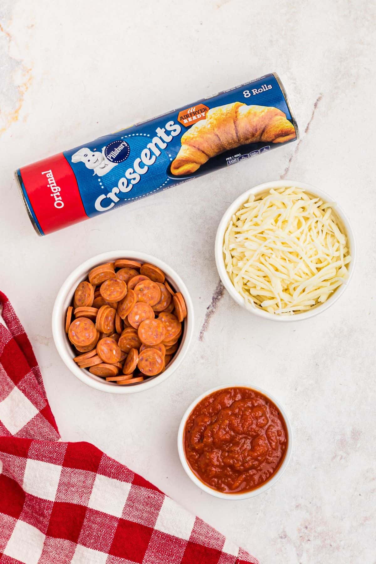 Can of crescent roll dough with small bowls of pepperoni, shredded mozzarella cheese, and pizza sauce. 