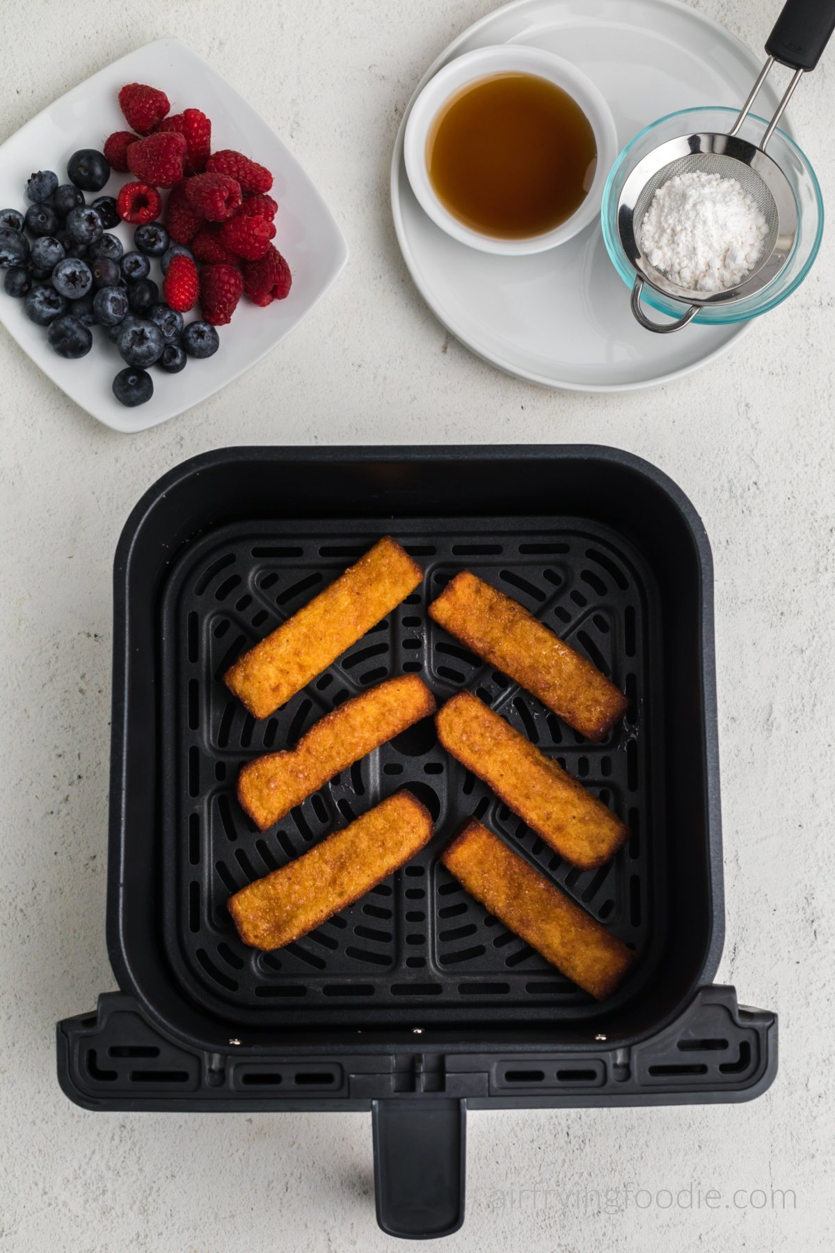 french toast sticks in air fryer basket, fully cooked from frozen. Berries, syrup, and powdered sugar above the basket.