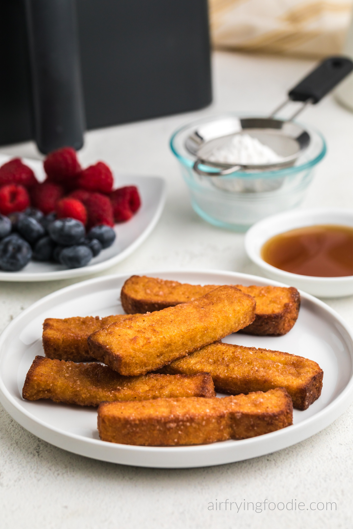 Air fried frozen french toast sticks, fully cooked and served on a white plate with berries, syrup, and powdered sugar in the background. 