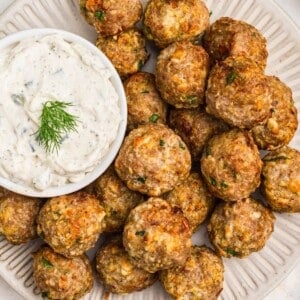 Golden Greek meatballs on a white plate with sauce next to it.