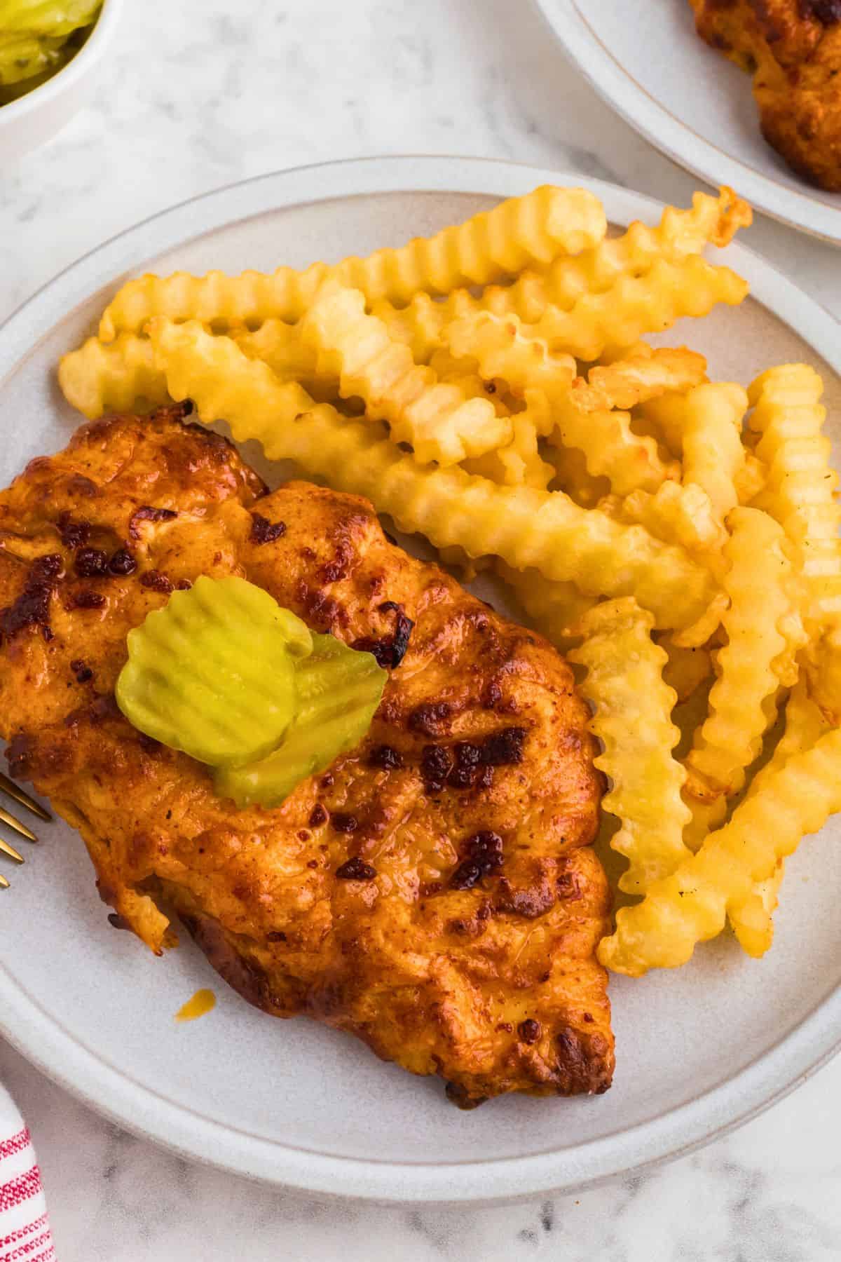 Juicy Nashville hot chicken on a white plate with a side of fries, topped with pickles. 