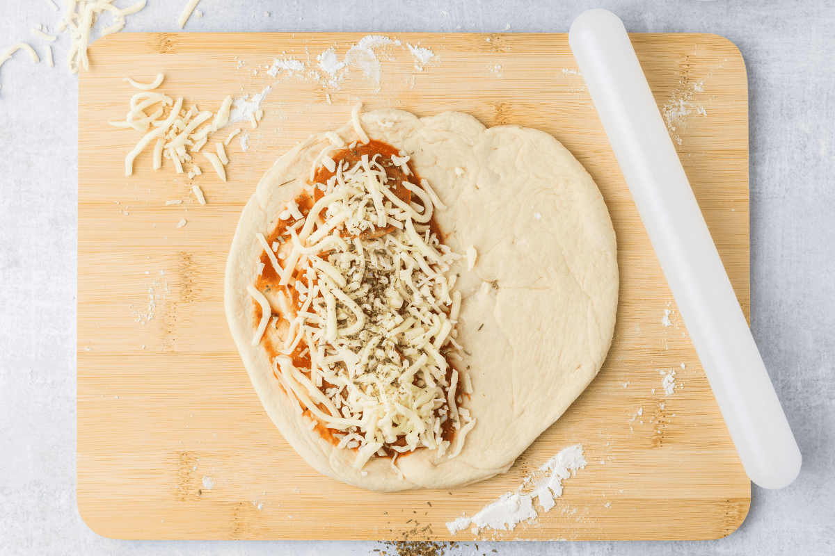 Dough rolled into a circle, then topped with sauce, cheese, pepperoini, and seasoning. On a cutting board. 